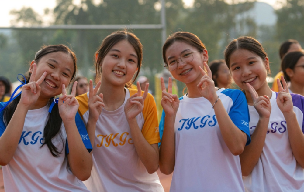 Four smiling TKGS students in colourful house shirts posing with peace signs during an outdoor school event.